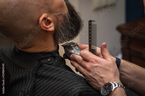 Close-up of a barber trimming a beard with electric clippers in a modern barbershop. Professional men's grooming and beard care service, selective focus