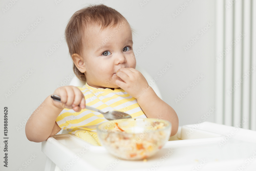 cute little baby eating, child sitting in high chair, holding spoon, good appetite