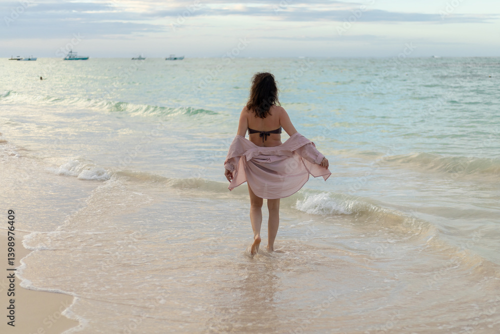 Woman walking toward the water on a beach during a calm evening with gentle waves and boats in the distance Woman walking toward the water on a beach during a calm evening with gentle waves and boats in the distance