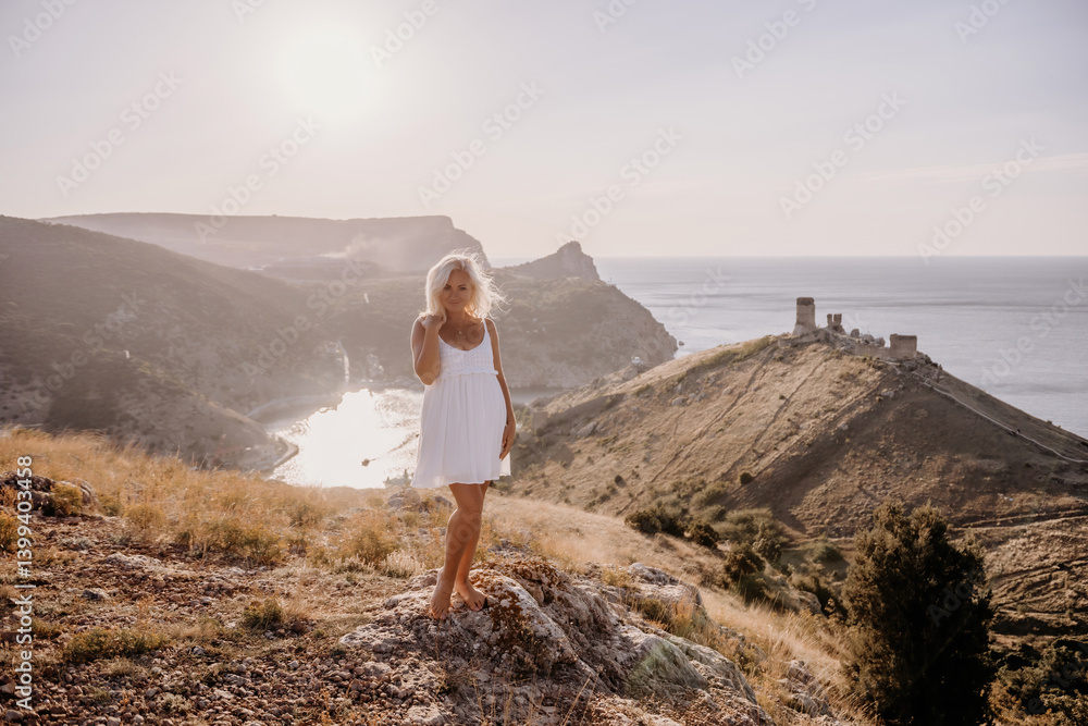 Naklejka premium A blonde woman stands on a hill overlooking the ocean. She is wearing a white dress and she is enjoying the view.