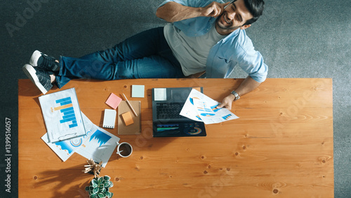 Relaxed caucasian businessman talking phone with team while put legs on table. Smart manager speaking and calling with colleague about marketing strategy while looking at financial graph. Convocation.