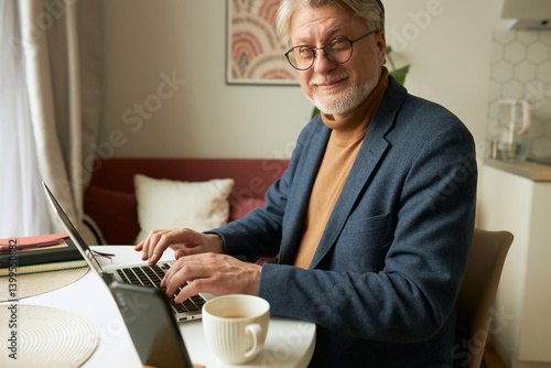 Side view of charming elderly intelligent elegant man author or writer in glasses working on laptop at table with coffee and smartphone at home office writing novel for children, inventing new plot