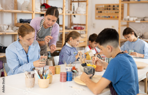 Children and teenagers sit at a table and paint ceramics at the workshop. Teacher shows how to paint a clay product after firing