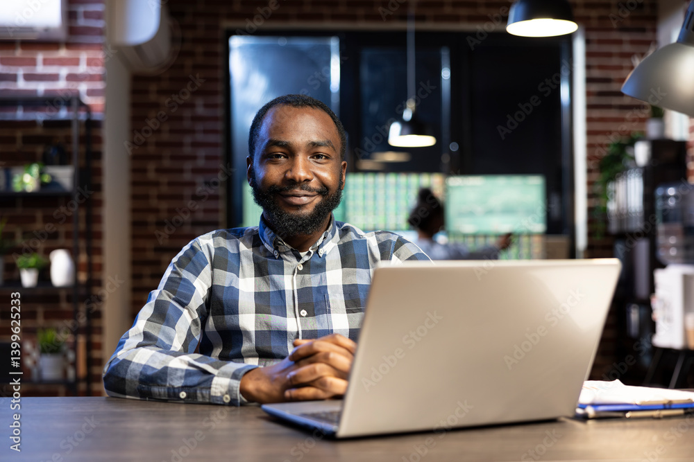 African american male professional sitting at a desk in a modern office with a brick wall, looking at the camera and smiling. His laptop and clipboard are set up for reviewing financial data. African american male professional sitting at a desk in a modern office with a brick wall, looking at the camera and smiling. His laptop and clipboard are set up for reviewing financial data.