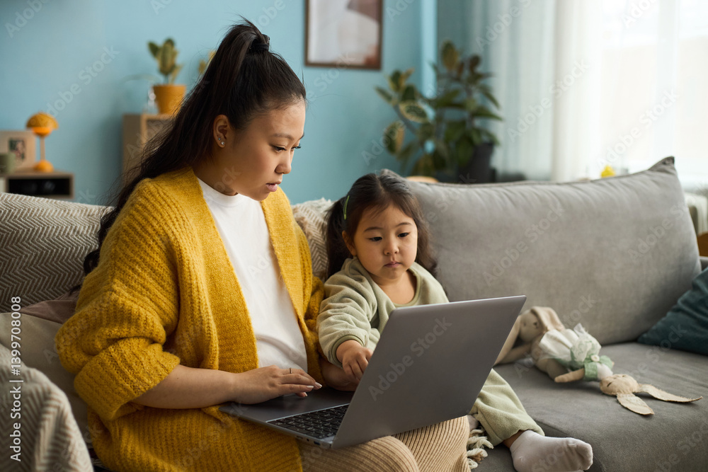 Fototapeta premium Caucasian mother and young daughter sitting on couch, using laptop together in cozy home environment, both focused and engaged without dot at the end
