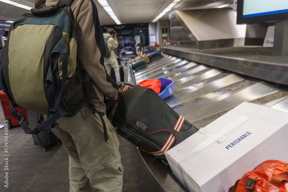 Man pulling suitcase from the luggage conveyor belt. Box with Perishable label on it. Unrecognizable people collecting their luggage at the airport.