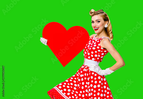 Pinup beautiful woman show red heart shape paper banner with mockup space. Pin up cute girl. Retro fashion vintage ad concept. Isolated over vibrant green chroma key background. Valentine or like sign