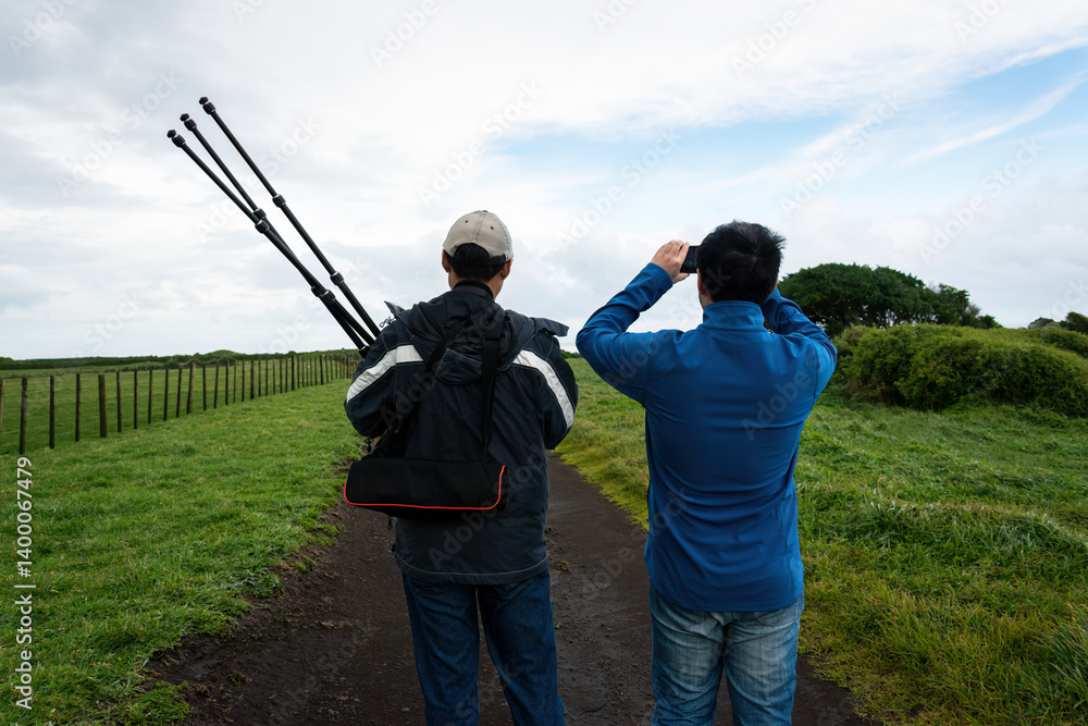 Two men walking on a rural road. One man taking smartphone photos. Another man holding tripod. Taranaki. New Zealand.