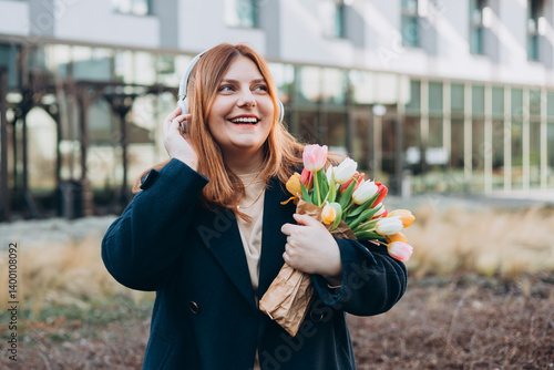 Obraz na płótnie Beautiful young happy woman in headphones with bouquet of tulip posing on urban background. Smiling 30s girl listening to music through wireless headphones w salonie