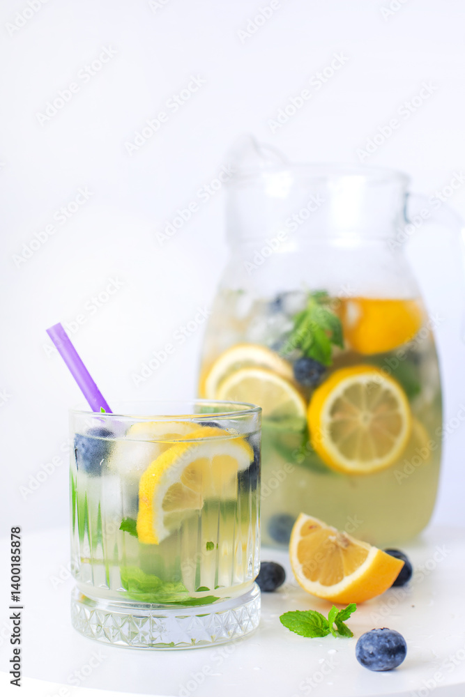 Fototapeta premium Chilled citrus drink with lemon slices, blueberries, mint, and ice in a glass and jug, on a white background.