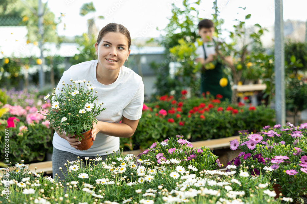 Smiling young girl choosing potted plants for flower garden in greenhouse in spring time, looking at white blooming daisies with interest