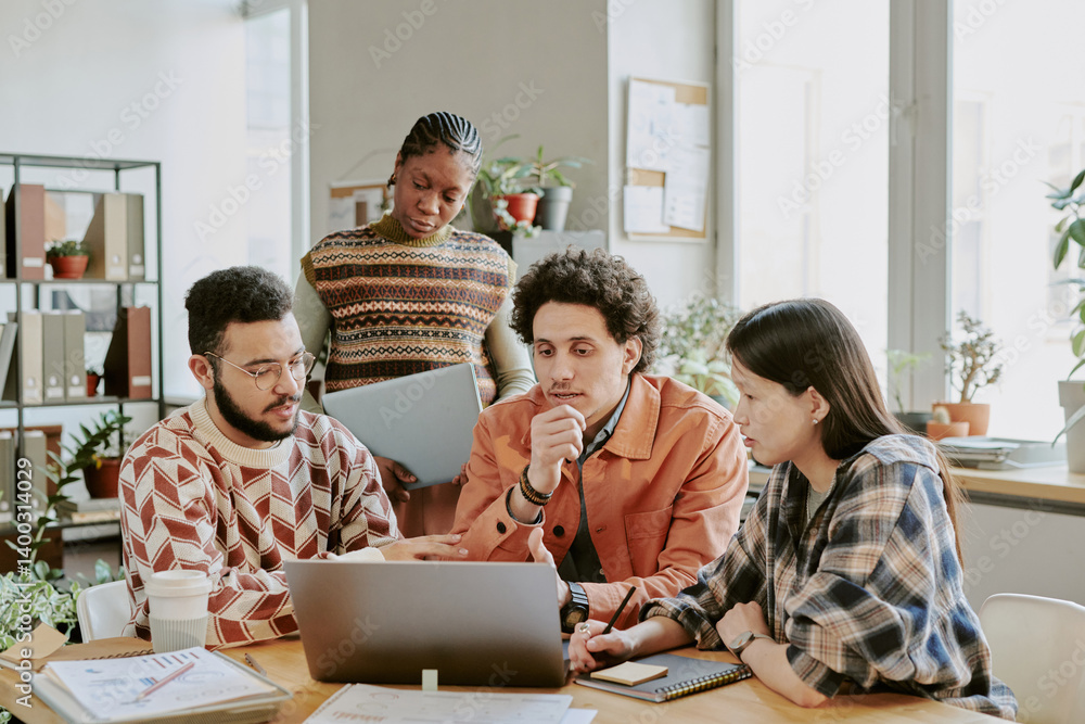 Medium shot of diverse programmers sitting at table looking at screen of laptop while working in collaborative and dynamic environment Medium shot of diverse programmers sitting at table looking at screen of laptop while working in collaborative and dynamic environment