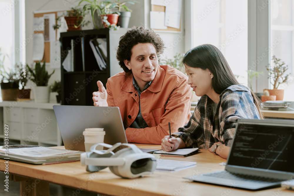 Biracial male and female IT specialists sitting at work desk together and having nice conversation during work day in office Biracial male and female IT specialists sitting at work desk together and having nice conversation during work day in office