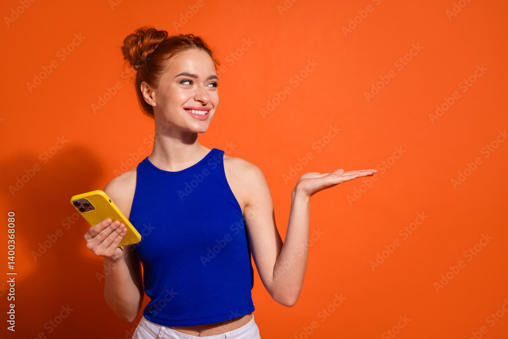 Smiling Young Woman Holding a Yellow Smartphone and Gesturing With Her Hand Against a Vibrant Orange Background Smiling Young Woman Holding a Yellow Smartphone and Gesturing With Her Hand Against a Vibrant Orange Background