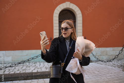 Young 30s woman in a black coat and sunglasses, smiling and posing for a selfie while holding a bouquet of flowers. She is standing outside in front of a vibrant orange wall, capturing a moment of joy