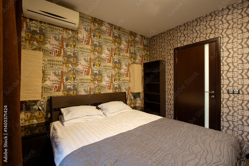 Apartment interior. Double bed in the bedroom. Pillows and bedspread on the made bed. Wallpaper with images of European landmarks. Air conditioning and wall lamps on the wall. Door in the wall.