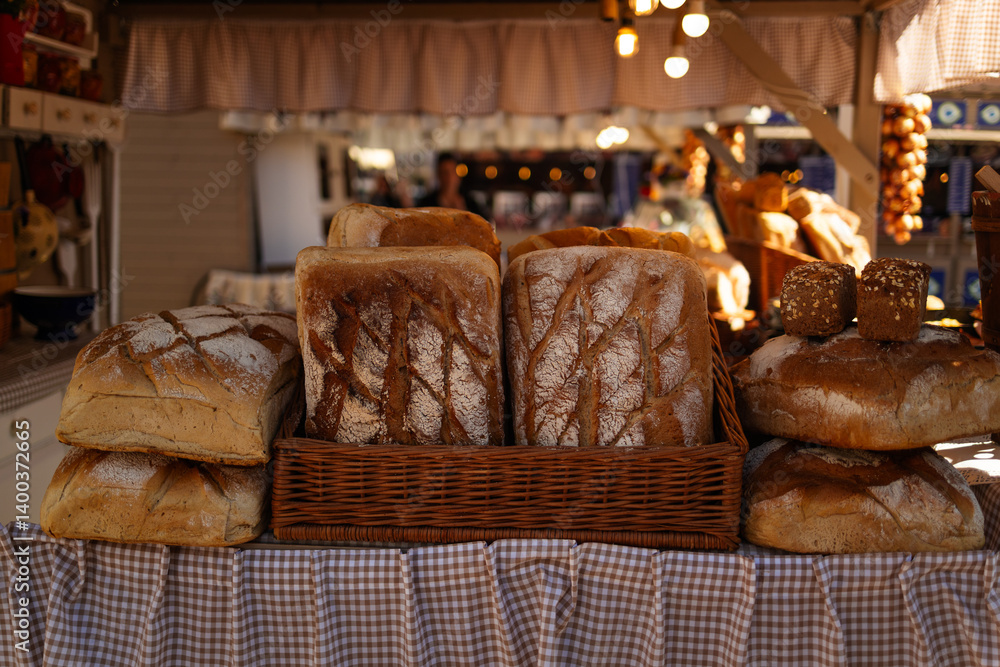 A variety of freshly baked loaves of bread are neatly arranged on a display table at a market. The loaves are golden-brown with crisp crusts and sit in a basket with a checkered cloth. Lights hang