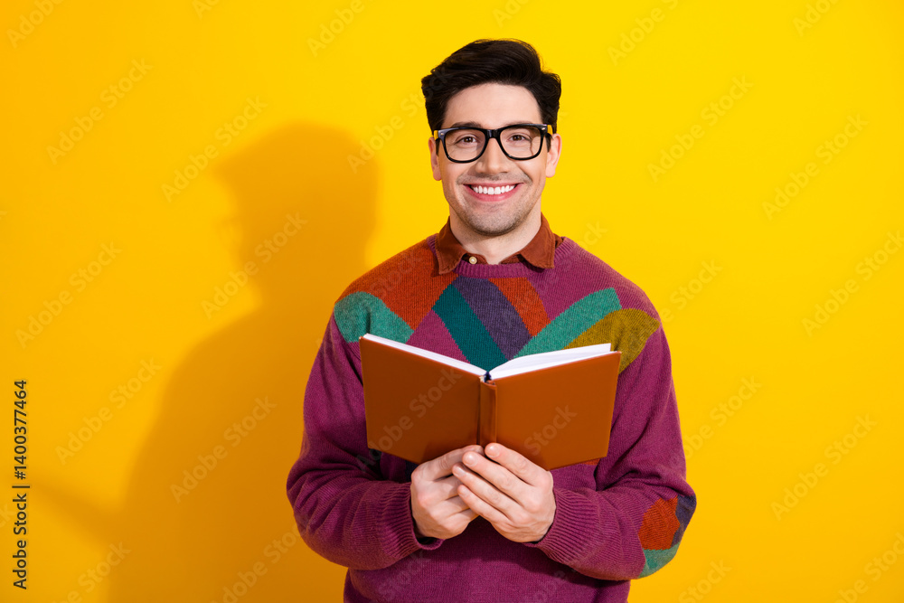 Smiling young man with glasses holding an open book in front of a vibrant yellow background, wearing a colorful sweater. Smiling young man with glasses holding an open book in front of a vibrant yellow background, wearing a colorful sweater.
