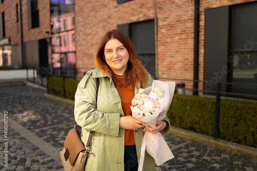 Obraz na płótnie A joyful woman smiles as she holds a bouquet of flowers in an urban setting with modern brick buildings and greenery in the background. She's dressed stylishly in a green coat and brown accessories. w salonie