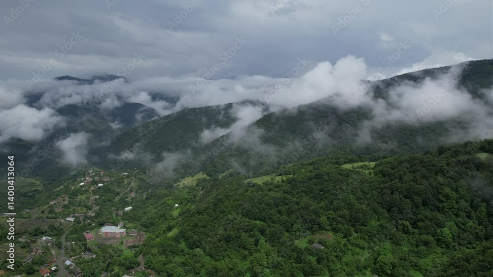 Scenic drone view of low clouds over a green lush mountain with scattered houses under cloudy sky