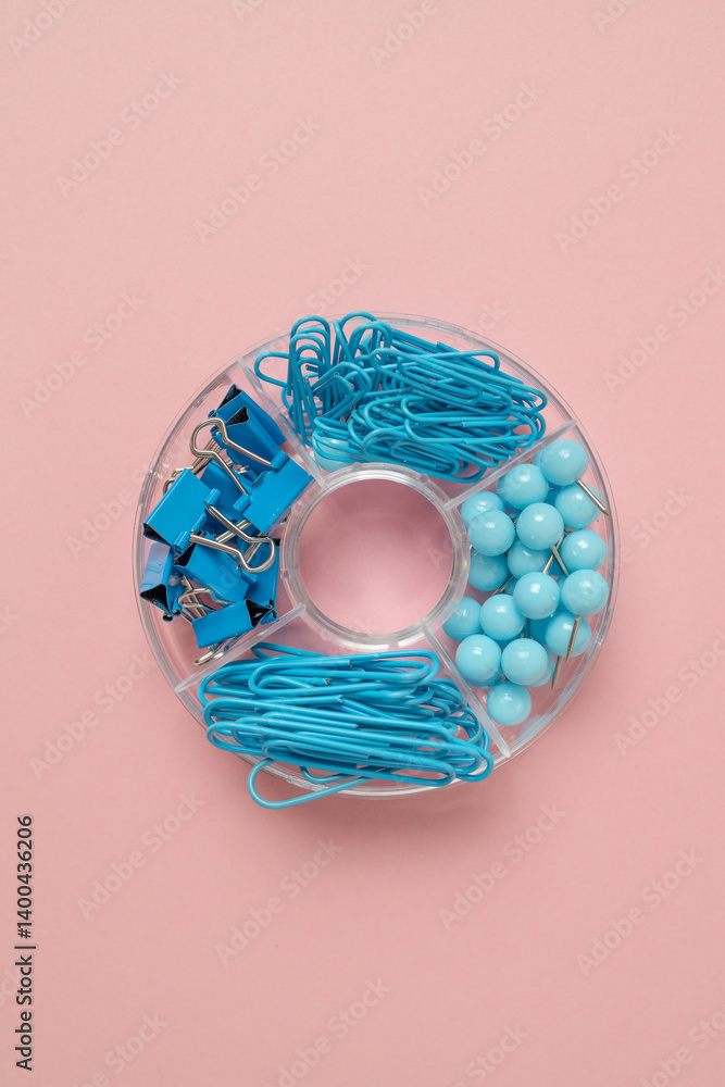 A creative arrangement of blue office supplies, including paper clips, rubber bands, and push pins, neatly organized in a circular container against a soft pink background. A colorful and stylish