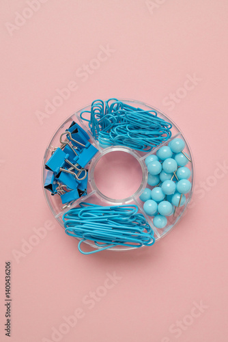 Obraz na płótnie A creative arrangement of blue office supplies, including paper clips, rubber bands, and push pins, neatly organized in a circular container against a soft pink background. A colorful and stylish w salonie