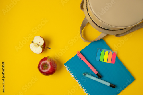 A vibrant and colorful setup featuring office supplies such as pens, sticky notes, and a notepad, complemented by a fresh red apple. The bright yellow background adds an energetic touch to the scene