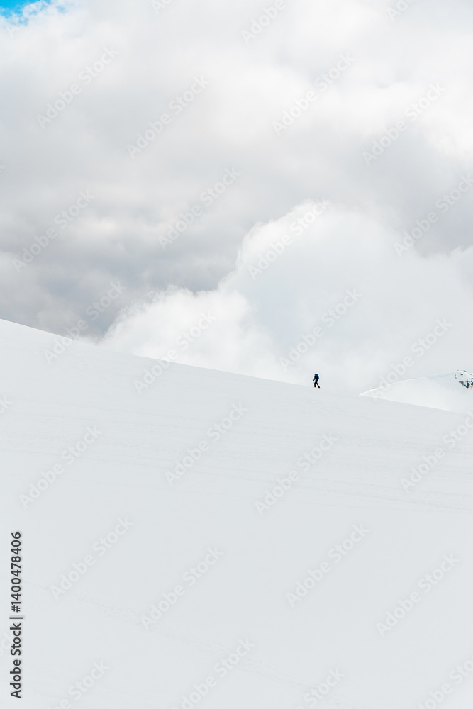 Lone hiker on a snow-covered slope under cloudy skies in Sierra de Gredos, Spain, minimalist vertical winter scene highlighting solitude and vast alpine scale Lone hiker on a snow-covered slope under cloudy skies in Sierra de Gredos, Spain, minimalist vertical winter scene highlighting solitude and vast alpine scale