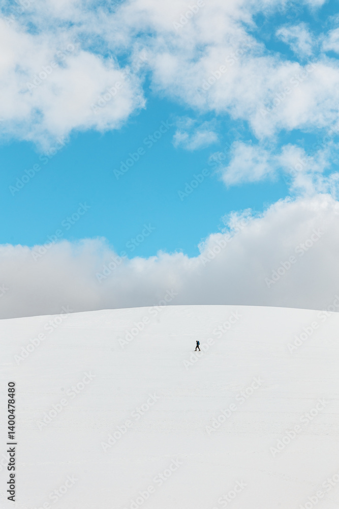 Lone hiker walking across a snow-covered hill beneath a vibrant blue sky in Sierra de Gredos, Spain, captured in a minimalist vertical winter landscape Lone hiker walking across a snow-covered hill beneath a vibrant blue sky in Sierra de Gredos, Spain, captured in a minimalist vertical winter landscape