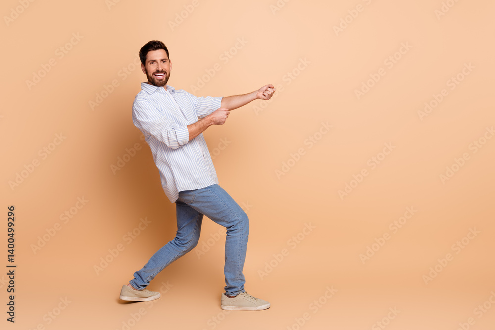 Smiling young man posing in a stylish casual outfit against a beige background with a playful gesture of pulling an imaginary rope. Smiling young man posing in a stylish casual outfit against a beige background with a playful gesture of pulling an imaginary rope.