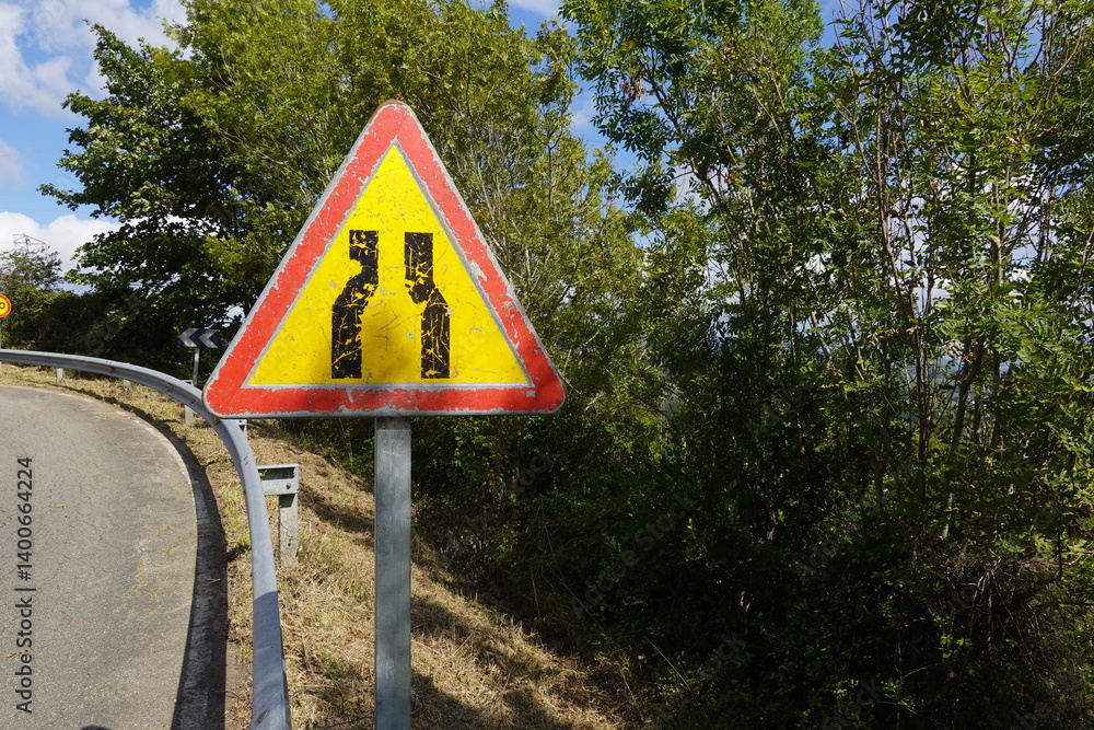 A worn warning sign showing road narrowing stands beside a rural road, surrounded by greenery and trees, with faded paint from exposure. A worn warning sign showing road narrowing stands beside a rural road, surrounded by greenery and trees, with faded paint from exposure.