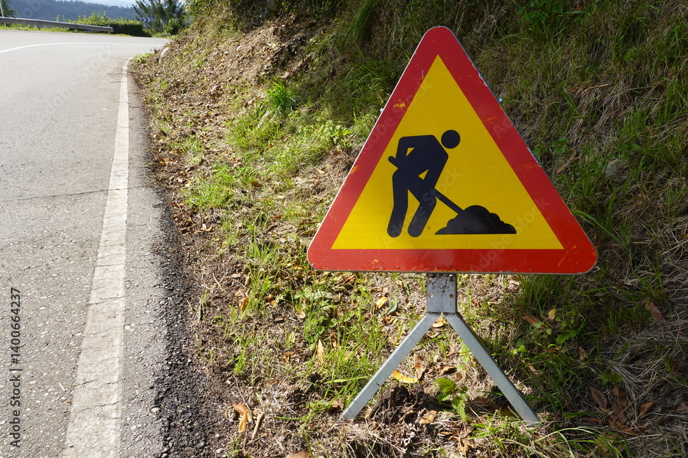 A roadwork warning sign showing a construction worker is placed along a rural road, with grass and greenery around the area. A roadwork warning sign showing a construction worker is placed along a rural road, with grass and greenery around the area.
