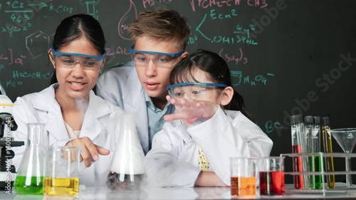 Girl looking at beaker while doing experiment at blackboard with chemistry theory in STEM class. teacher and group of high school student with mixed races doing science activity together. Edification