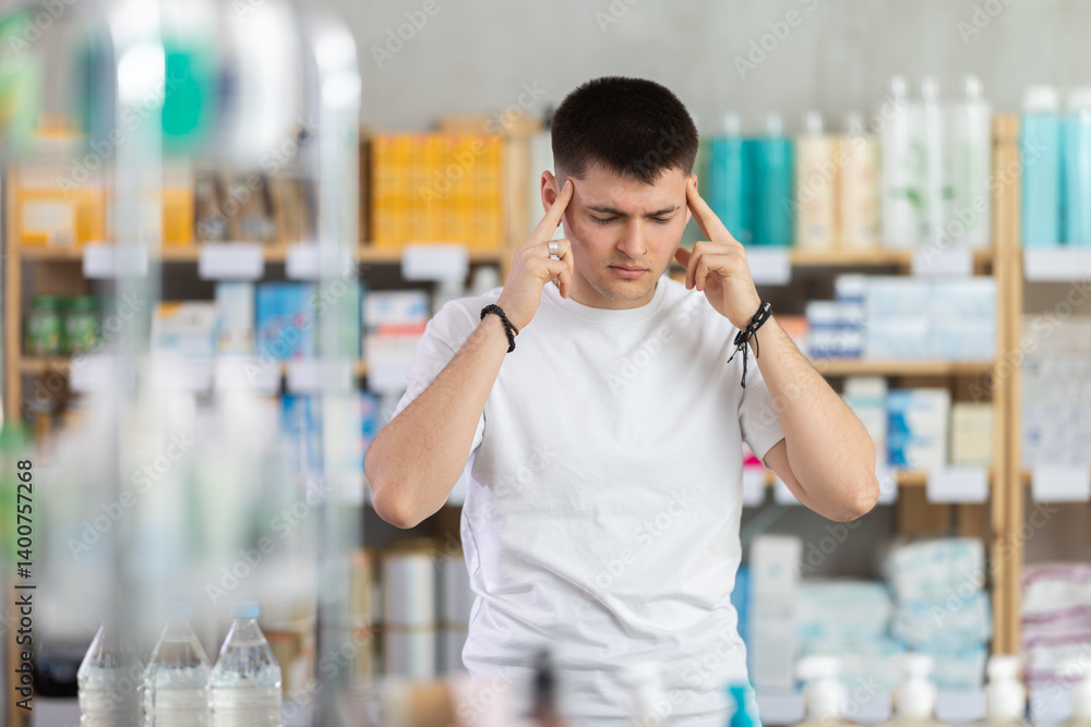 Sick man standing between shelves in small drugstore suffering from headache or having migraine, choosing medicine. Health care and medicine concept Sick man standing between shelves in small drugstore suffering from headache or having migraine, choosing medicine. Health care and medicine concept