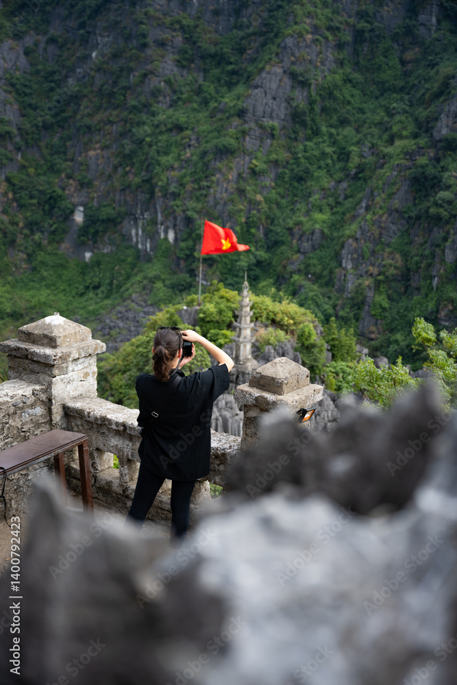 (Selective focus) A girl is taking pictures from the Hang Múa Viewpoint with a pagoda and the Vietnamese national flag in the distance. Ninh Binh, Vietnam.
