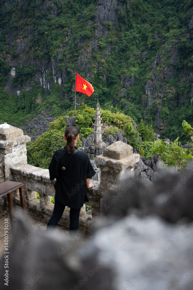 (Selective focus) Defocused girl enjoying the view from the Hang Múa Viewpoint with a pagoda and the Vietnamese national flag in the distance. Ninh Binh, Vietnam.