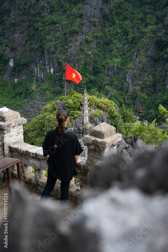 Obraz na płótnie (Selective focus) Defocused girl enjoying the view from the Hang Múa Viewpoint with a pagoda and the Vietnamese national flag in the distance. Ninh Binh, Vietnam. w salonie