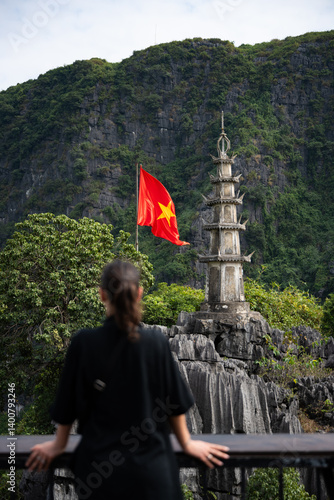 Obraz na płótnie (Selective focus) Defocused girl enjoying the view from the Hang Múa Viewpoint with a pagoda and the Vietnamese national flag in the distance. Ninh Binh, Vietnam. w salonie