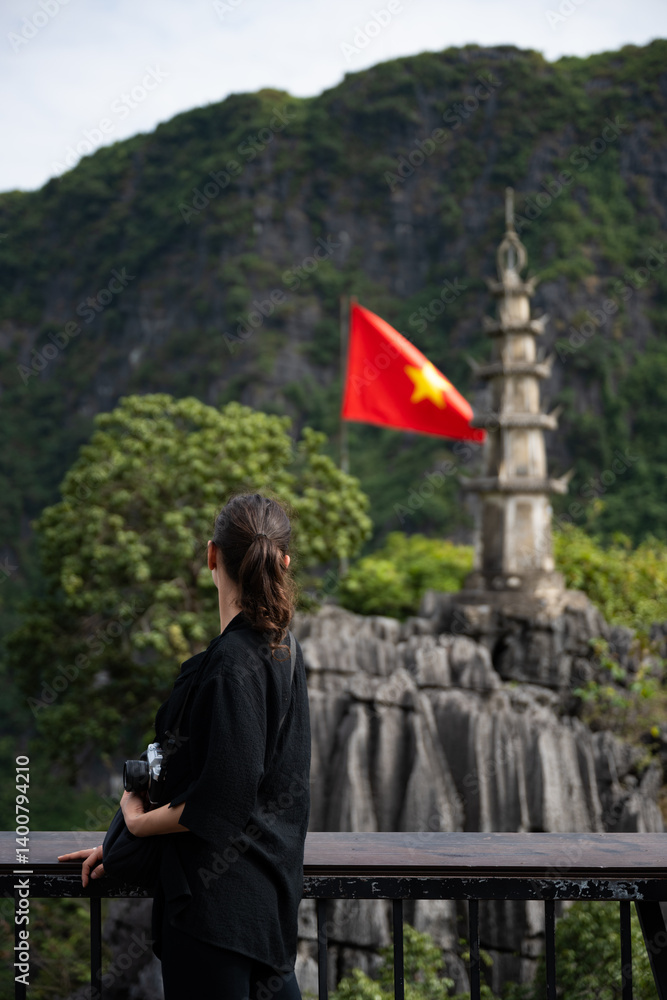 (Selective focus) Stunning view of a girl enjoying the view from the Hang Múa Viewpoint with a pagoda and the Vietnamese national flag in the distance. Ninh Binh, Vietnam.