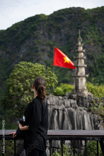 Obraz na płótnie (Selective focus) Stunning view of a girl enjoying the view from the Hang Múa Viewpoint with a pagoda and the Vietnamese national flag in the distance. Ninh Binh, Vietnam. w salonie