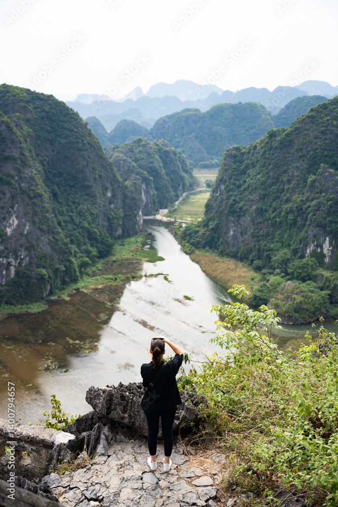 (Selective focus) A girl is taking pictures from the Hang Múa Viewpoint with the a stunning landscape and the Tam Coc river in the distance. Ninh Binh, Vietnam.