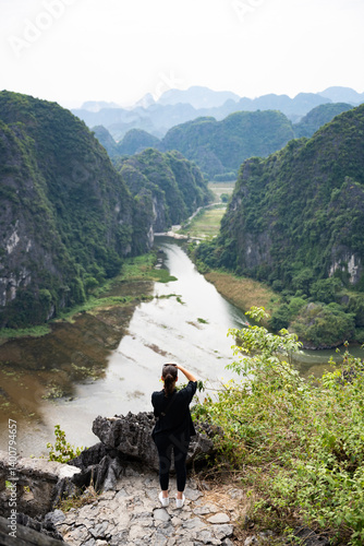 Obraz na płótnie (Selective focus) A girl is taking pictures from the Hang Múa Viewpoint with the a stunning landscape and the Tam Coc river in the distance. Ninh Binh, Vietnam. w salonie