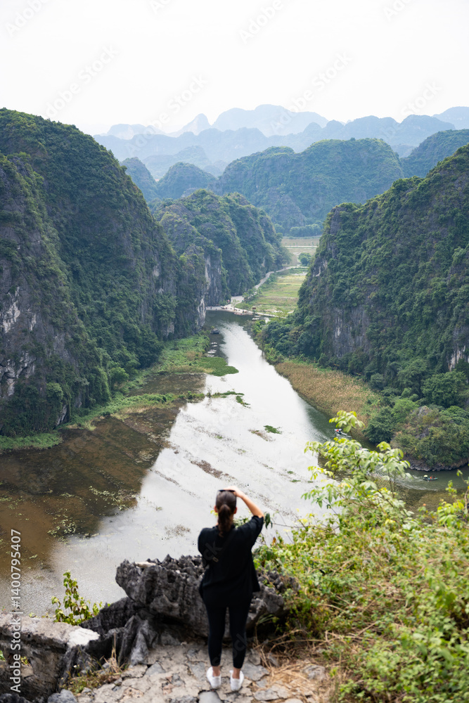 (Selective focus) Defocused girl taking pictures from the Hang Múa Viewpoint with the a stunning landscape and the Tam Coc river in the distance. Ninh Binh, Vietnam.