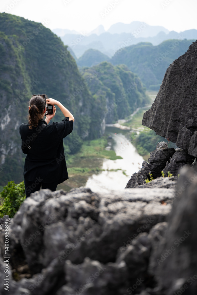 (Selective focus) A girl is taking pictures from the Hang Múa Viewpoint with the a stunning landscape and the Tam Coc river in the distance. Ninh Binh, Vietnam.