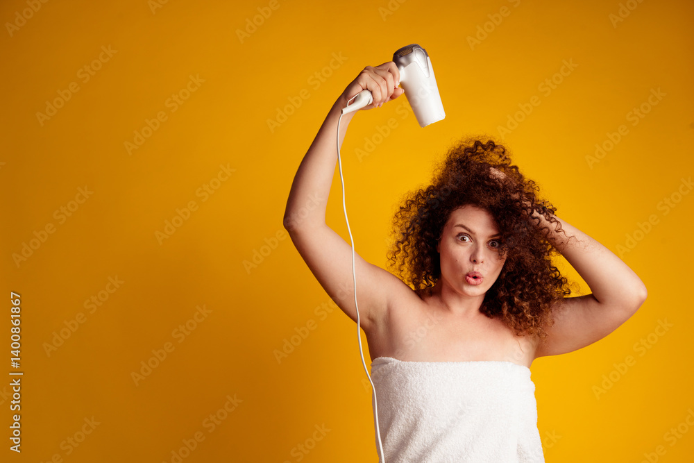 Portrait of a young woman drying her curly hair with a blow dryer against a bright yellow background Portrait of a young woman drying her curly hair with a blow dryer against a bright yellow background