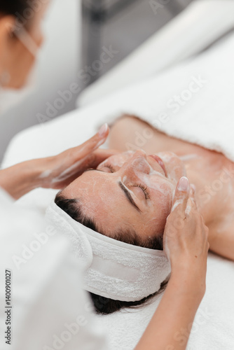 Cosmetologist doing female face massage with creamy foam, skin beauty treatment, improving appearance. Relaxed girl getting professional facial rejuvenating, cleansing, moisturizing in beauty clinic