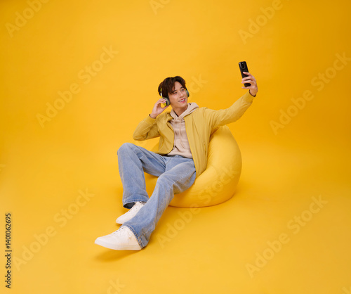 Happy young asian man listening to music with headphone and using phone sitting chair isolated on yellow background. People lifestyle concept.