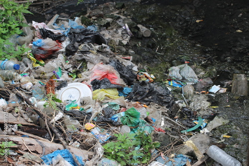 A powerful environmental image showing a polluted riverbank filled with plastic waste, bags, bottles, and other garbage