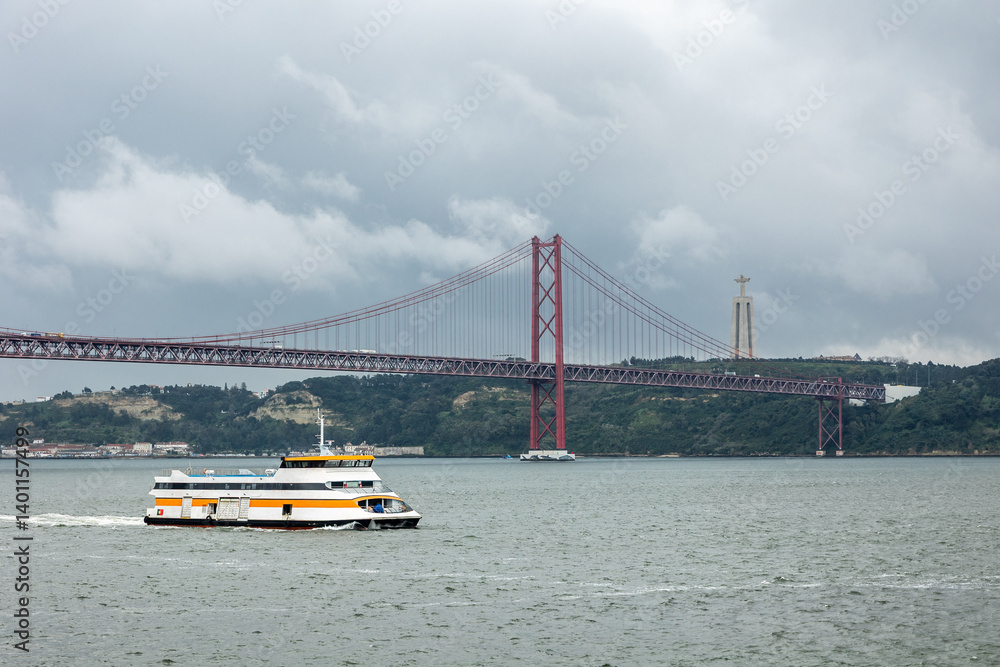 Fototapeta premium A scenic ferry crosses the Tagus River in Lisbon, Portugal, with the iconic 25 de Abril Bridge and Cristo Rei statue in the background under a dramatic cloudy sky.