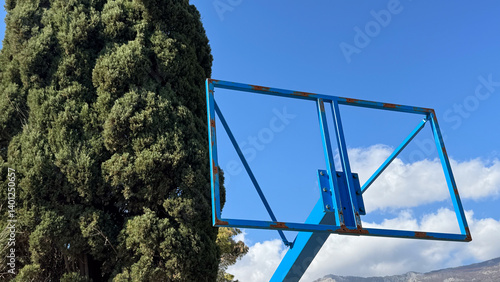 Obraz na płótnie Rusty, hollow blue basketball hoop frame against tall evergreen tree and blue sky.
w salonie